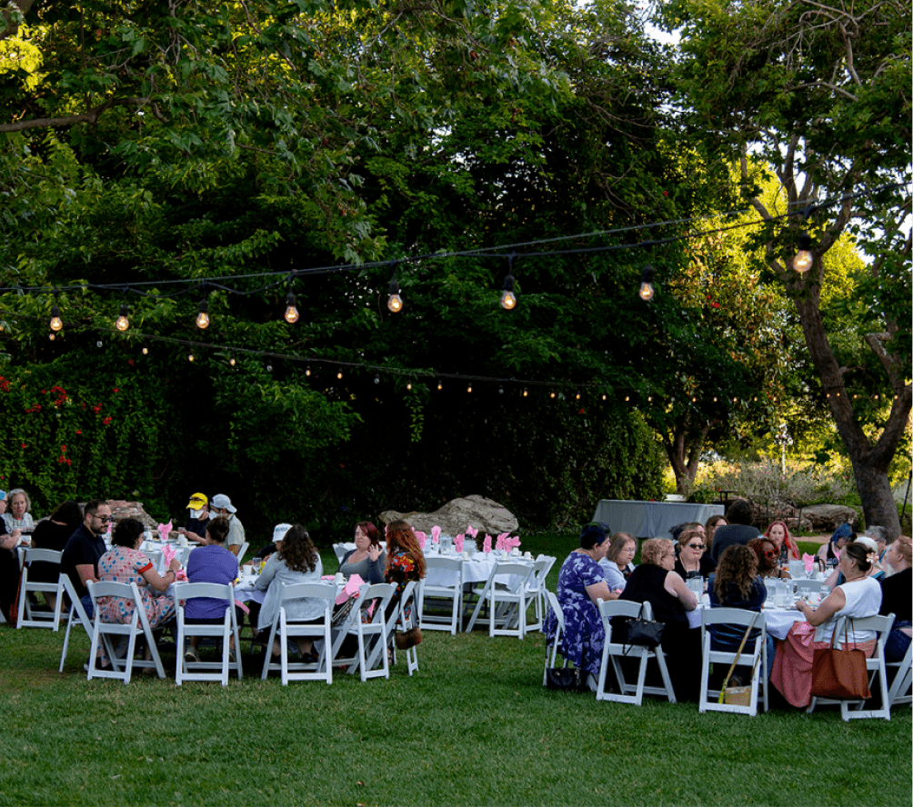 Guests seated at round tables under string lights during an outdoor dinner event in a garden setting.