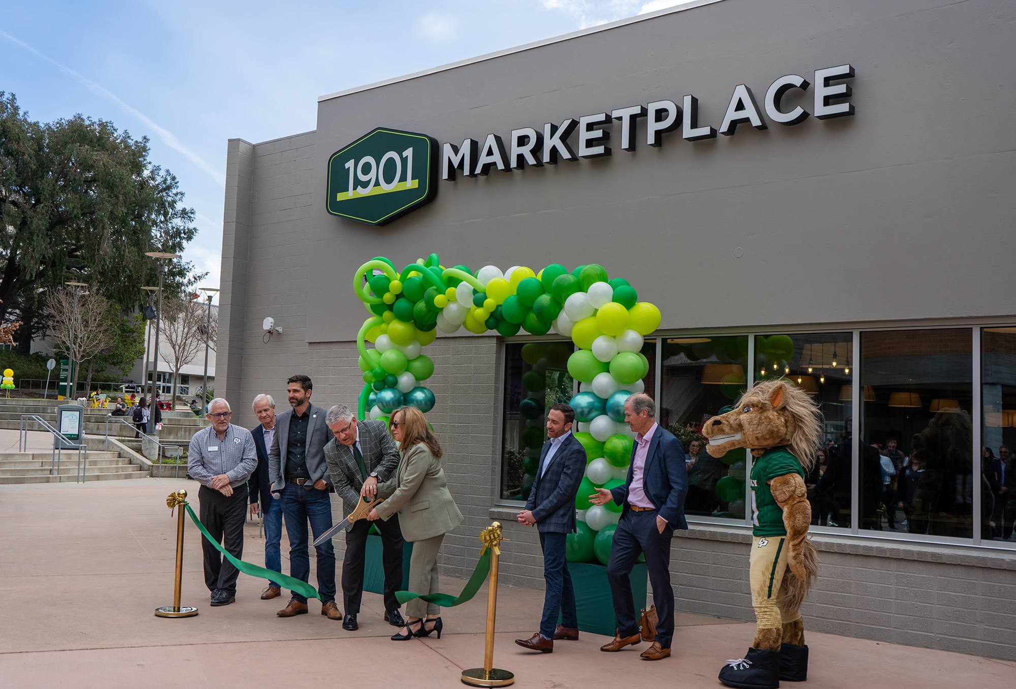 Ribbon-cutting ceremony for the 1901 Marketplace at Cal Poly, with university officials, green and white balloon arch, and mascot present.