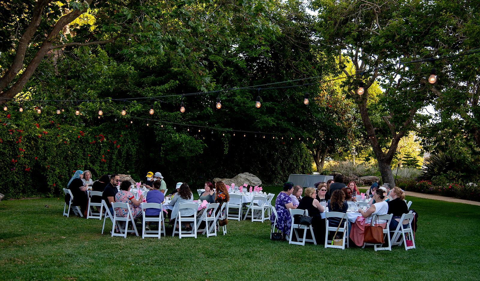 Guests seated at round tables during an outdoor garden dinner under string lights, surrounded by lush greenery.