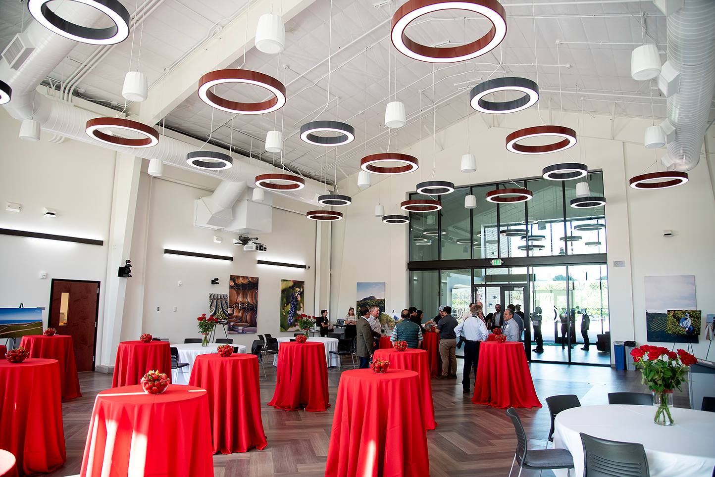 Wide-angle view of a modern event space with circular ring lights on the ceiling, red tablecloth-covered cocktail tables with bowls of strawberries, and guests networking near the glass entrance.