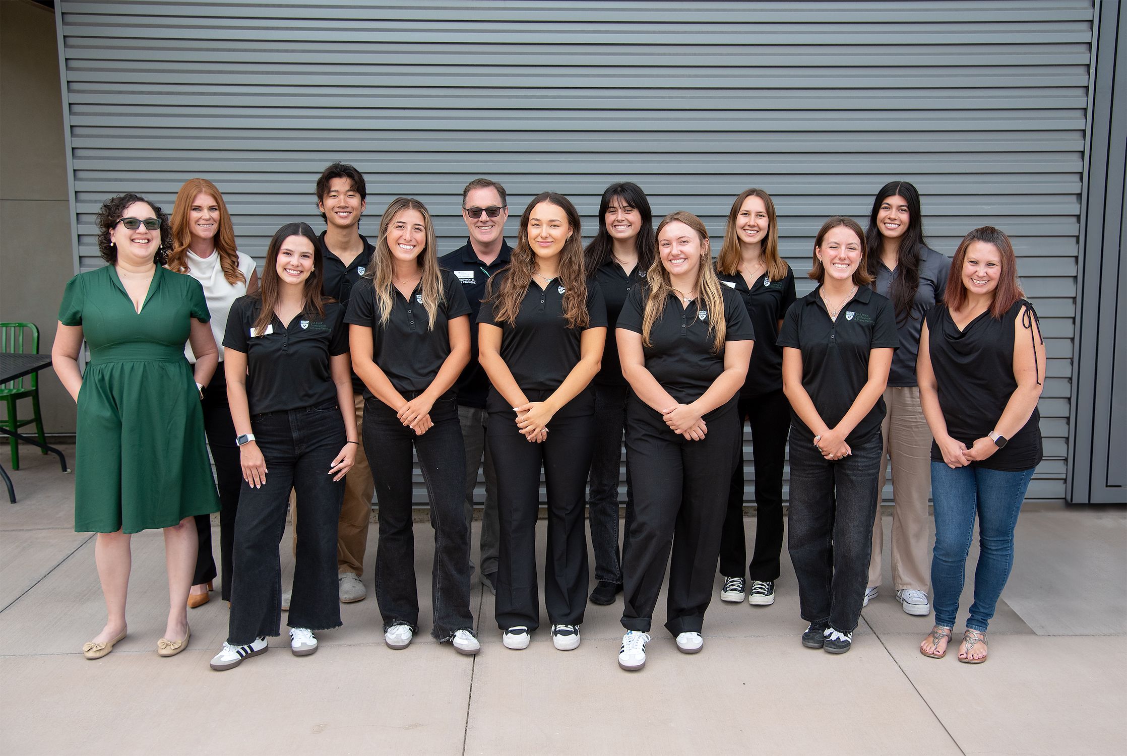 Group photo of a diverse team of students and staff standing in two rows in front of a gray metal wall, with most wearing black Cal Poly attire and smiling at the camera.