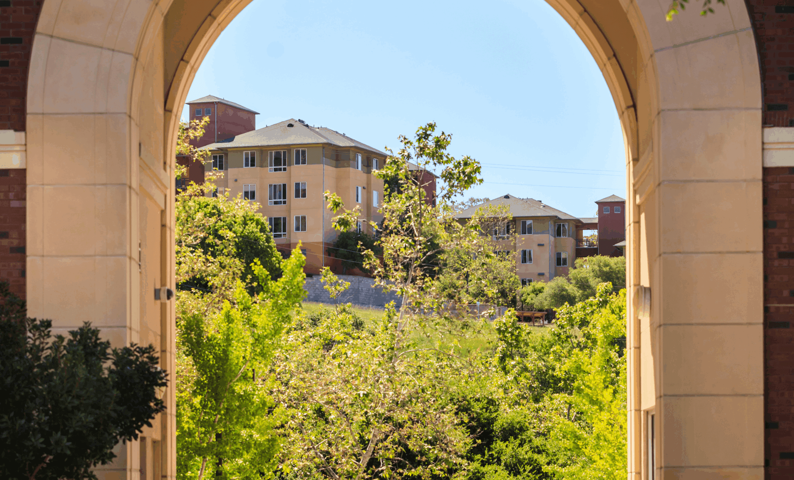 View through a large stone archway framing a cluster of hillside apartment buildings surrounded by greenery on a sunny day.
