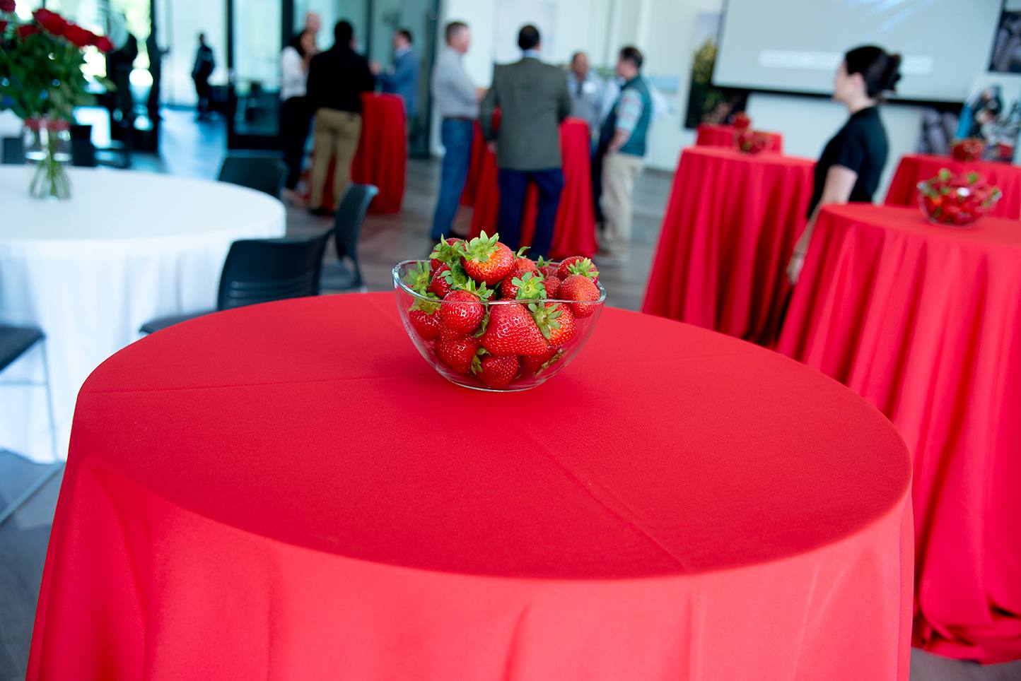 Clear glass bowl filled with fresh strawberries on a red tablecloth, with people mingling in the background at a networking event.